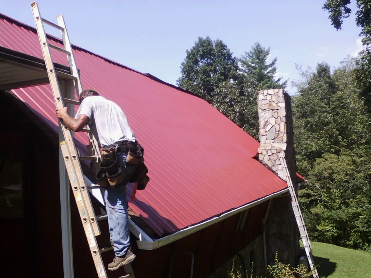 Roofer on a ladder beside a red metal roof and stone chimney — Soffit & Fascia Repair by Roofing Brothers in Waite Park